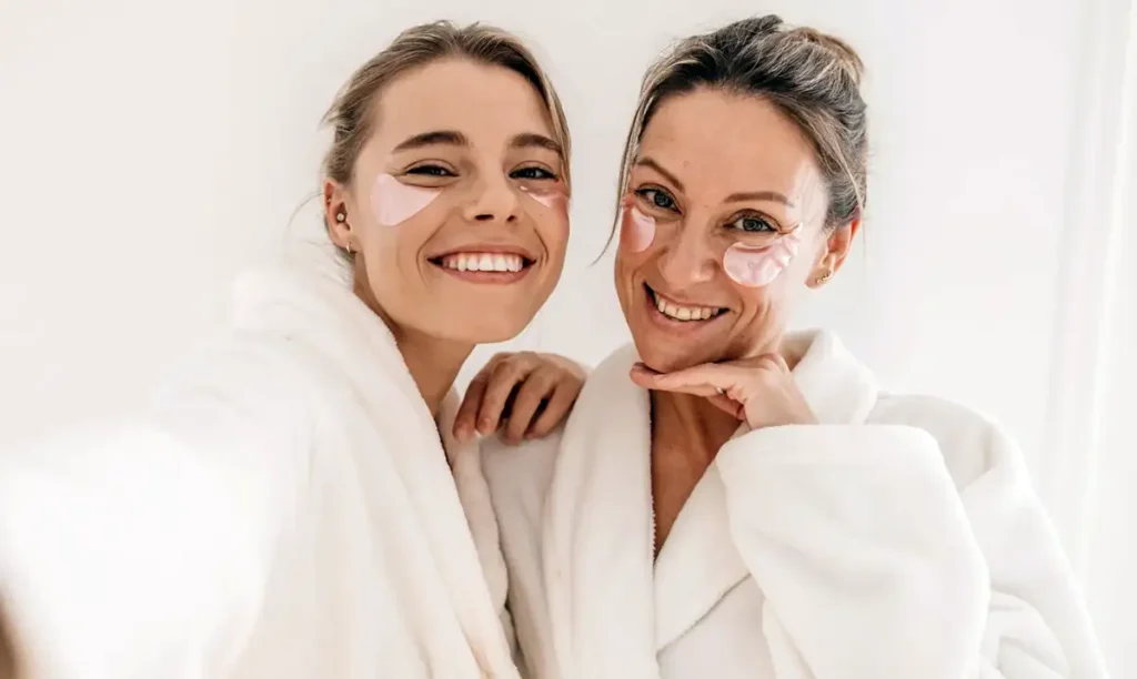 Deux femmes d'âges différents complices et souriantes, appliquant des patchs pour les yeux dans le cadre de leur routine après soin esthétique.